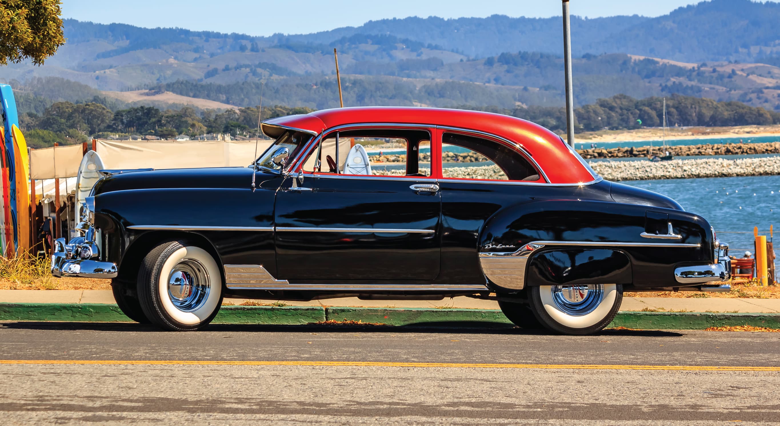 A vintage black sedan parked along the Half Moon Bay coastline with surfboards, ocean, and rolling hills in the background